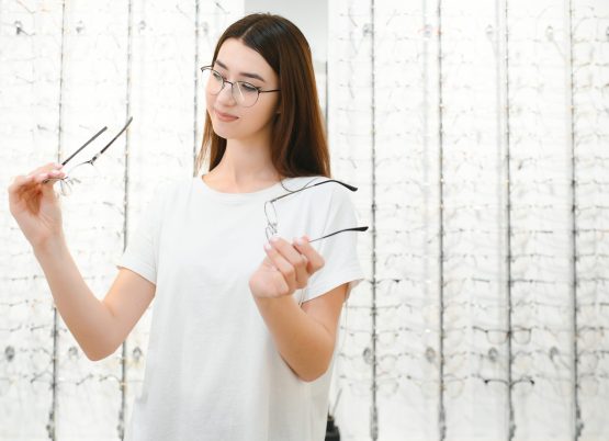 eyesight and vision concept - young woman choosing glasses at optics store